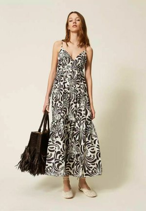Donna che indossa un abito lungo senza maniche a motivo bianco e nero, che tiene in mano una grande borsa tote nera intrecciata, in piedi su uno sfondo neutro.