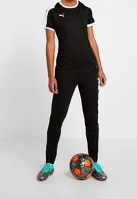 Zwarte sportshirt met witte accenten, gecombineerd met zwarte broek. Groene en grijze voetbalschoenen en een zwart en oranje voetbal maken de look compleet.