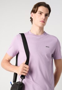 Camiseta de manga corta color lavanda con cuello redondo y un pequeño logo negro "BOSS" en el pecho, combinada con un bolso de hombro negro.