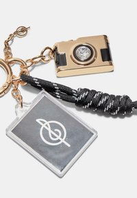 Ciondolo a forma di fotocamera in oro con un dettaglio in cristallo trasparente, accenti neri e un cordino bianco e nero. Include un'etichetta trasparente con un logo bianco.