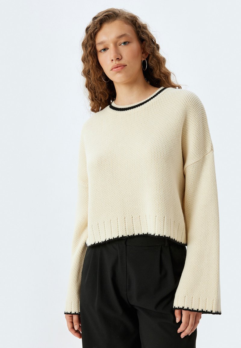 Maglione corporeo color crema realizzato in tessuto a maglia testurizzato, con bordi neri sul collo e sull'orlo, e maniche ampie per una vestibilità rilassata.