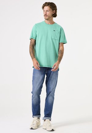 Camiseta de algodón verde con escote redondo y detalle de bolsillo, combinada con jeans de mezclilla azul y zapatillas beige. Ajuste casual con mangas cortas.