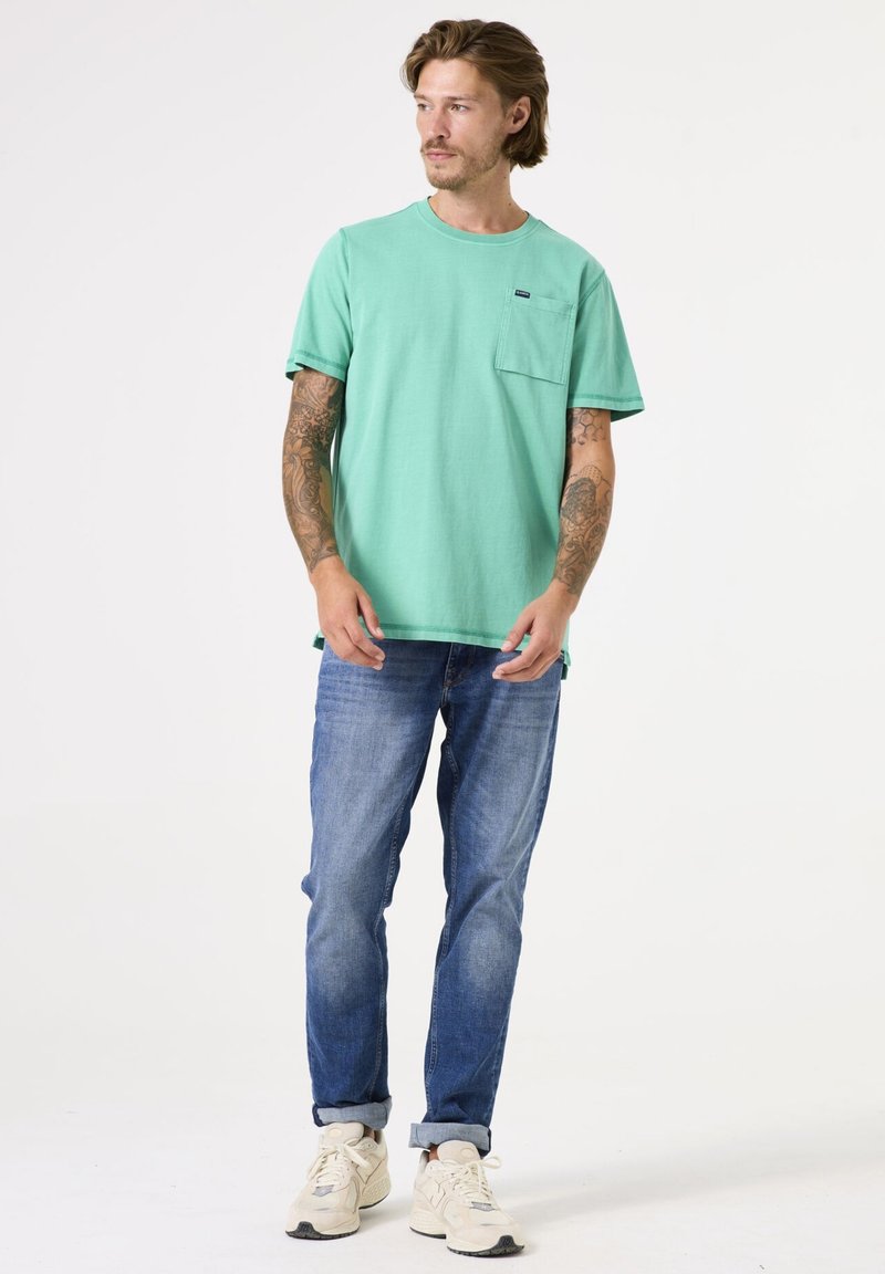 Groene katoenen T-shirt met een ronde hals en zakdetail, gedragen met blauwe spijkerbroek en beige sneakers. Casual fit met korte mouwen.