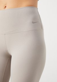 Beżowe legginsy z wysokim stanem wykonane z elastycznego materiału o gładkiej fakturze i minimalnych szwach, z subtelnym czarnym logo Nike.