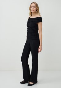 Femme portant un haut noir texturé à épaules dénudées et un pantalon évasé assorti, se tenant devant un fond clair, chaussée de ballerines noires.