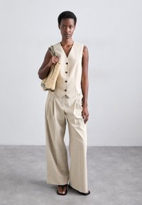Beżowa dopasowana kamizelka z guzikami w zestawieniu z szerokimi spodniami. Model trzyma fakturowaną beżową torbę, stojąc na neutralnej powierzchni.