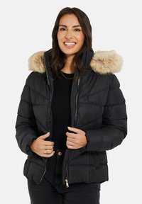 Zwarte pufferjas met een nepbontkap, ritssluiting aan de voorkant, quiltontwerp en een getailleerde pasvorm, gecombineerd met een zwarte top en donkere jeans.
