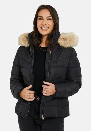 Zwarte pufferjas met een nepbontkap, ritssluiting aan de voorkant, quiltontwerp en een getailleerde pasvorm, gecombineerd met een zwarte top en donkere jeans.