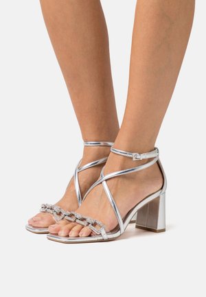 Sandales à talons hauts argentées avec un talon bloc, dotées de brides croisées à la cheville et d'une chaîne décorative sur le bout. Finition métallique lisse.