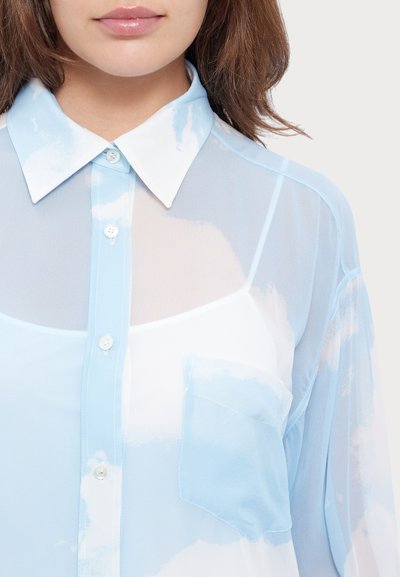 Chemisier léger bleu clair avec un col blanc, présentant des motifs ressemblant à des nuages, fermeture par boutons à l'avant et une poche unique sur la poitrine. Texture lisse.
