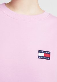 Ljusrosa sweatshirt med ribbad rundhals. Har en rektangulär Tommy Jeans-logomärke med blå, vit och röd accent på bröstet.
