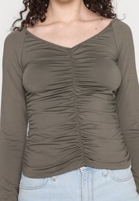 Olivgrünes Langarmoberteil mit gerüschtem Design an der Vorderseite und den Seiten, aus glattem, dehnbarem Material gefertigt.