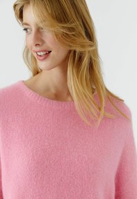 Junge Frau mit blondem Haar, die lächelt und nach links schaut, und einen weichen rosa Strickpullover trägt vor einem schlichten weißen Hintergrund.