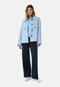 Hellblaues Wolljackett mit Kragen, Fronttaschen und schwarzen Knöpfen, kombiniert mit einem hellblauen Oberteil und dunkler Jeans. Weiße Sneakers.