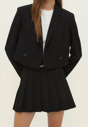 Kobieta ubrana w krótką czarną marynarkę z guzikami na białej bluzce, w połączeniu z czarną plisowaną spódnicą z wysokim stanem.