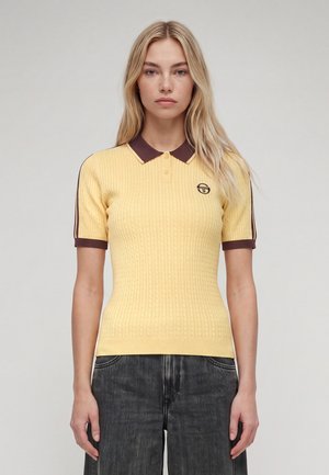 Jeune femme aux longs cheveux blonds portant un polo en maille jaune ajusté avec col et manches marron, et un jean gris foncé, debout devant un fond uni.