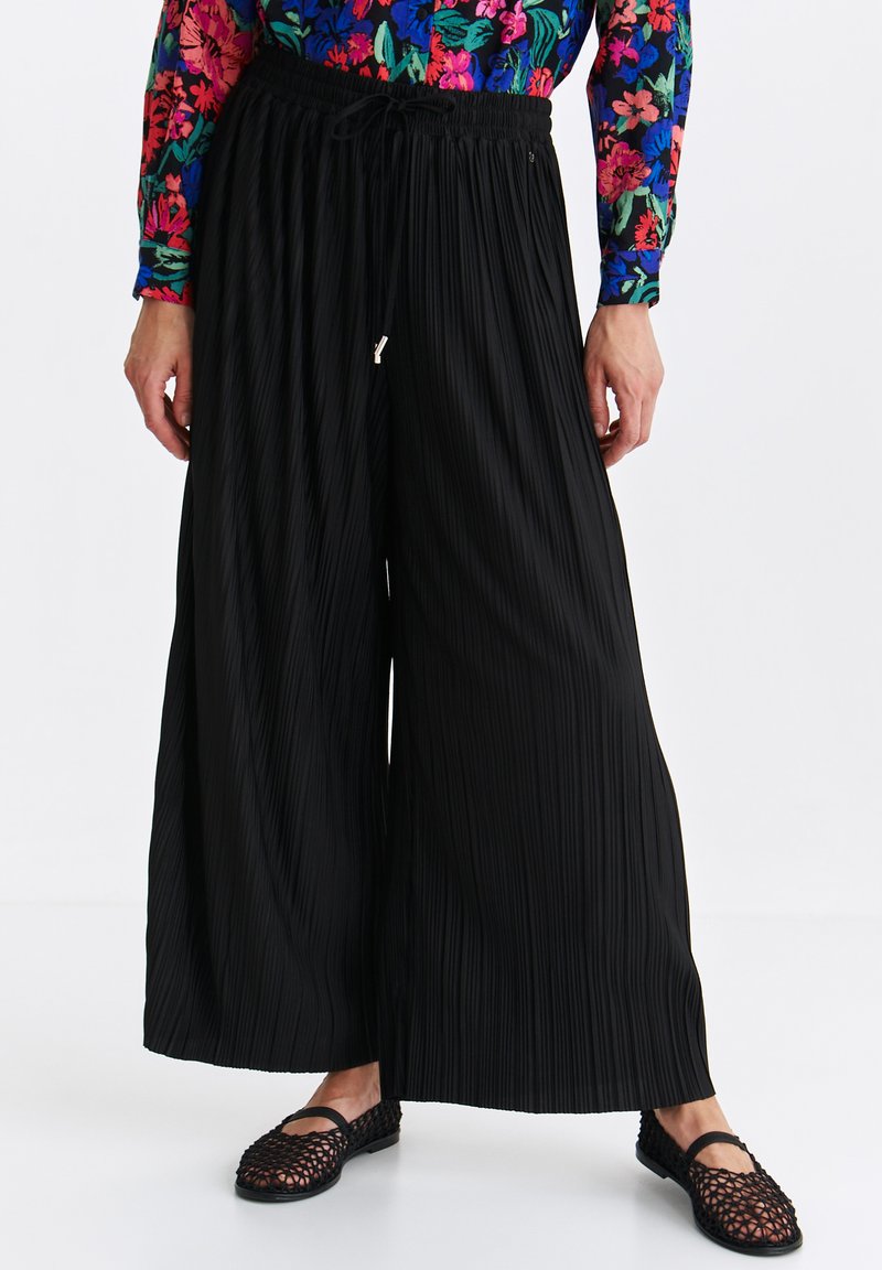 Czarne szerokie spodnie plisowane z elastycznym pasem i sznurkiem, połączone z teksturowanym czarnym obuwiem.