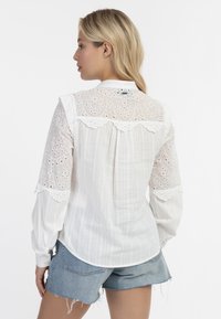 Witte blouse met ingewikkelde kantdetails op de schouders, zachte textuur, knoopsluiting bij de hals, gecombineerd met denim shorts.