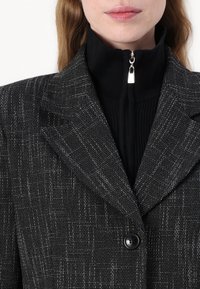 Černý blazer s texturovaným vzorem s jemnými kostkami. Vybaven širokými klopami, jedním knoflíkem a kontrastním černým zapínacím tričkem pod ním.