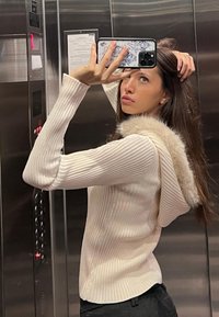 Maglione a costine crema con cappuccio foderato in pelliccia, caratterizzato da maniche lunghe e una silhouette aderente, indossato in un contesto di ascensore.