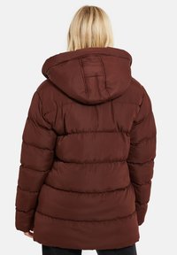 Personne blonde portant une doudoune marron avec une capuche, vue de dos sur un fond clair uni.