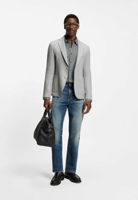 Grijze blazer over een patroonhemd, gecombineerd met blauwe jeans. Zwarte leren tas en schoenen. Soepele textuur, op maat gemaakt, casual uitstraling.
