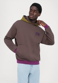 Felpa con cappuccio marrone con rivestimento interno verde, polsini viola e tasca frontale. Tessuto liscio con una vestibilità rilassata e dettagli di texture cuciti.