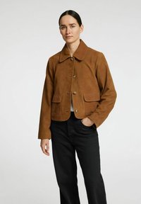 Brun ruskindsjakke med krave, to forlommer og knaplukning, båret med sorte jeans. Glat tekstur og kort design.