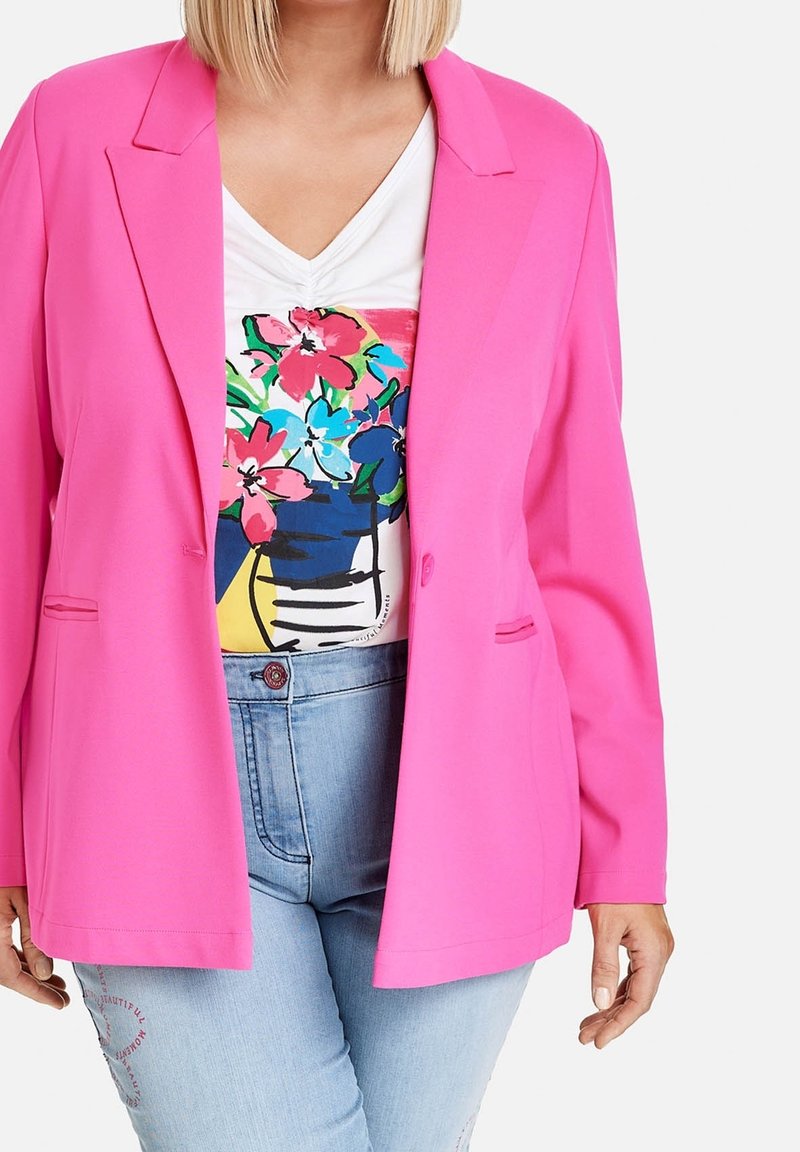 Kobieta w jasnoróżowej marynarce, białej koszulce z kwiatowym nadrukiem oraz jasnoniebieskich dżinsach z czerwonym haftem na udzie.