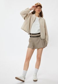 Chaqueta bomber cortada beige sobre una camiseta blanca, combinada con pantalones cortos a cuadros. Zapatillas blancas y una gorra marrón completan el look.