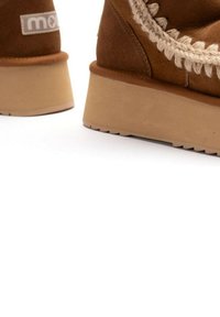 Stivali in suede marrone con tacco a zeppa, caratterizzati da dettagli intrecciati lungo la parte superiore e suole in gomma testurizzata per una migliore aderenza. Logo visibile sul lato.