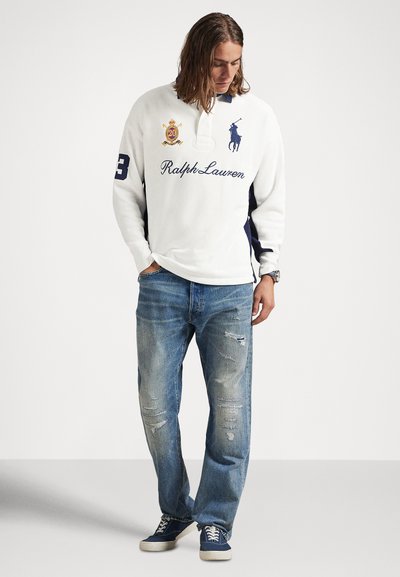 Νέος άνδρας με μακριά μαλλιά φορώντας λευκό πόλο Ralph Lauren με μακριά μανίκια, σκισμένα μπλε τζιν και μπλε αθλητικά παπούτσια, κοιτάζοντας προς τα κάτω.