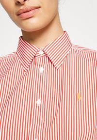 Red and white striped button-up shirt with a pointed collar and a small gold logo on the chest, made from cotton fabric.