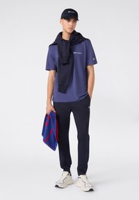 Navy t-shirt met logo, navy sweatshirt over de schouders, navy broek, houdt een rode en blauwe handdoek vast, beige sneakers. Eenvoudig sportief ontwerp.