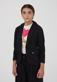 Blazer nero con collo a scialle e maniche arricciate, con un accento argento. Indossato sopra una camicia bianca con grafiche rosa e nere.