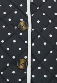 Quiltet jakke med svart bunn og hvite prikker, knappelukkinger og kontrastfarget hvit kant. Strukturert stoff med diamantmønster.