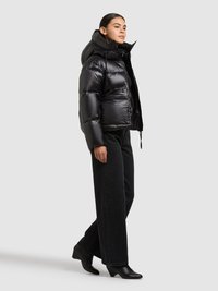 Zwarte donzen jas met een hoge kraag en capuchon, met een glanzende afwerking, ritssluiting en zijzakken, gedragen met zwarte broek met wijde pijpen.