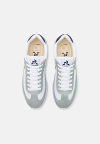 Zapatillas de ante gris claro con detalles en cuero blanco, punta redonda, cordones blancos y talón azul marino. Logo en la lengüeta y en la plantilla.