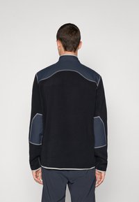 Black fleece jacket featuring blue shoulder and elbow patches, a stand collar, and contrasting gray stitching. Smooth texture and relaxed fit.