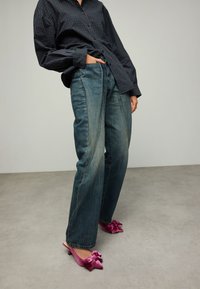 Jean bleu taille haute à jambes larges avec une finition délavée, associé à des mules en velours rose ornées de nœuds. Le modèle pose avec une main dans la poche.