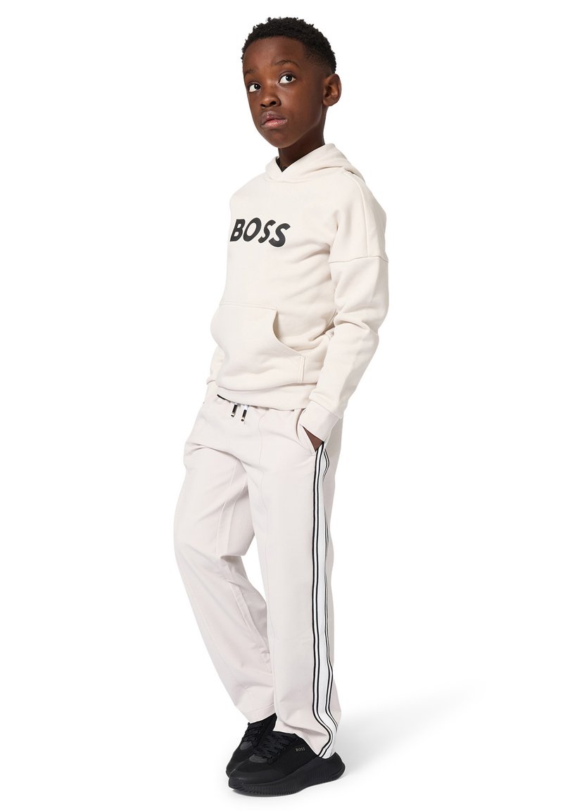 Garçon portant un sweat à capuche crème avec le texte "BOSS", un pantalon de survêtement assorti avec des bandes latérales noires, et des baskets noires, debout les mains dans les poches.