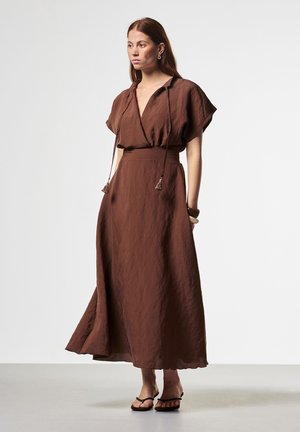 Vrouw draagt een bruine, korte mouw, enkelhoge jurk met een V-hals en tassel koordjes, gecombineerd met zwarte sandalen en een armband.