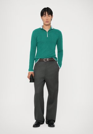 Grøn zip-neck sweater med tekstureret stof, parret med grå bredbenede bukser. Sort bælte og lille sort accesorium i hånden.