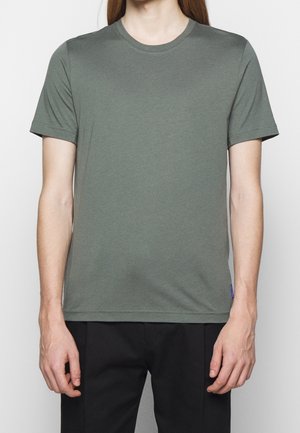 Green cotton crew neck t-shirt with short sleeves, smooth texture, and a small blue tag on the side. Worn with black trousers.