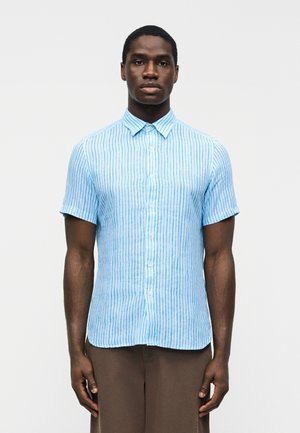 Άνδρας που φορά κοντομάνικο, γαλάζιο πουκάμισο με κάθετες λευκές ρίγες και καφέ παντελόνι, στέκεται μπροστά σε απλό φόντο.