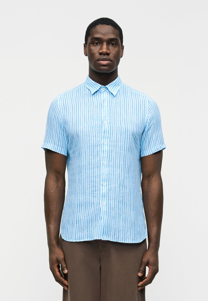 Man wearing a short-sleeve, light blue button-up shirt with vertical white stripes and brown pants, standing against a plain background.