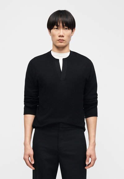 Νέος άνδρας που φοράει μαύρο μακρυμάνικο πουκάμισο με βαθύ V λαιμόκοψη πάνω σε λευκό T-shirt με στρογγυλή λαιμόκοψη, στέκεται μπροστά σε απλό ανοιχτό φόντο.