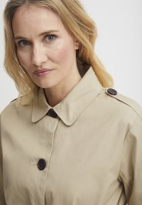 Beiger Trenchcoat mit klassischem Kragen, Knopfverschluss und glatter Baumwollstruktur. Mit dunklen Knöpfen an den Schultern.