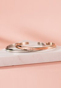 Trois bracelets jonc en métal dans des tons argent, or rose et or, présentant une finition lisse et brillante avec le nom de la marque gravé sur chacun.