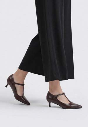 Persona que lleva pantalones negros de pierna ancha y zapatos de tacón bajo tipo kitten heel con punta puntiaguda y tira en T marrón oscuro sobre un fondo claro.